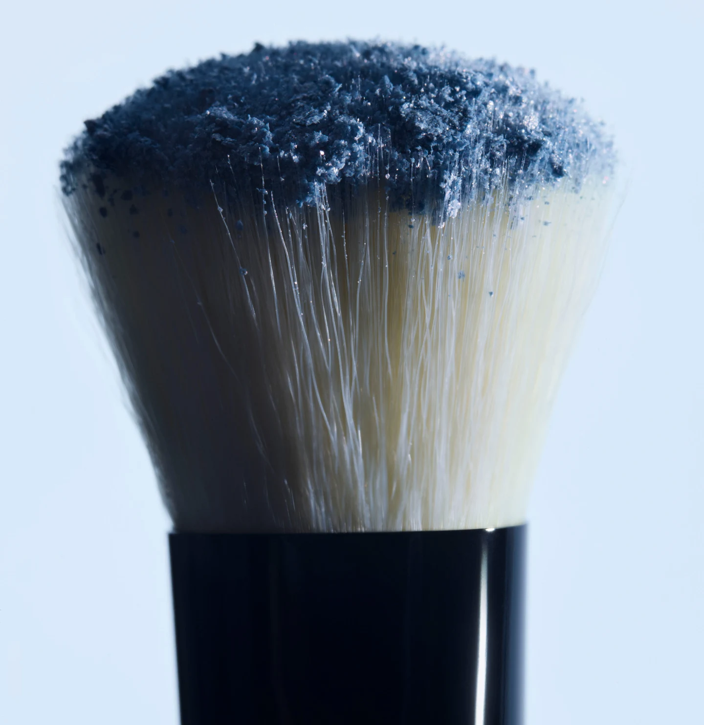 Extreme close-up of a makeup brush dipped in textured denim blue pigment, demonstrating the sheer wash of color technique for a quiet luxury look
