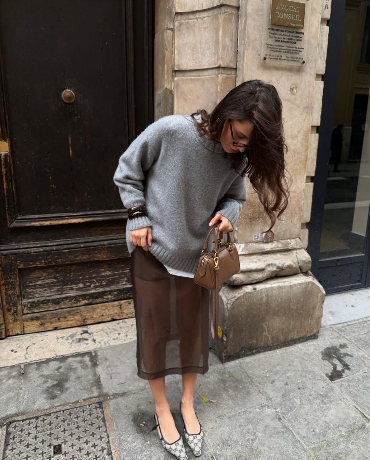 A sophisticated quiet luxury outfit featuring an oversized grey cashmere sweater paired with a sheer brown maxi skirt and high heels, demonstrating texture layering for